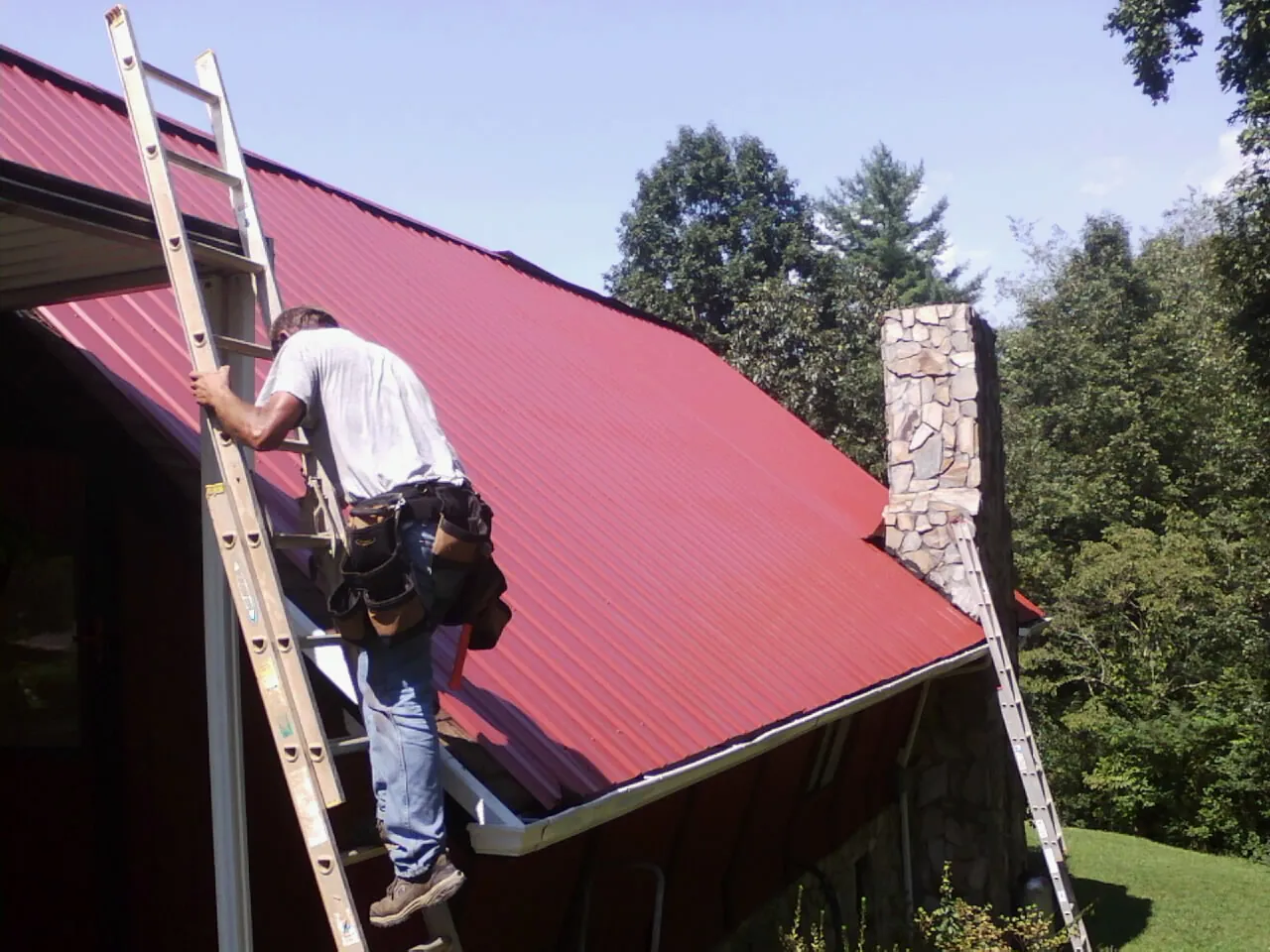 Roofer on a ladder beside a red metal roof and stone chimney — Soffit & Fascia Repair by Roofing Brothers in Massena